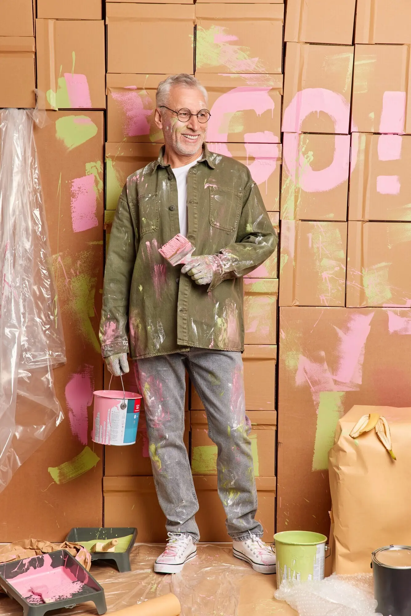 Fröhlicher Senior hält einen Farbeimer und einen Pinsel, führt Reparaturen durch, blickt erfreut zur Seite, trägt legere Kleidung und ist zu Hause mit der Renovierung beschäftigt. Konzept: Umbau, Hausverbesserung, Instandhaltung.